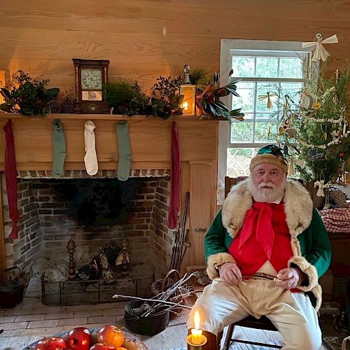 A man dressed as Santa Claus sits in a festive room with a fireplace, stockings, a Christmas tree, and holiday decorations, holding a musical instrument.