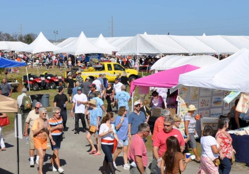 People are walking around a bustling outdoor market with many white tents and stalls displaying various items and a yellow vehicle visible.