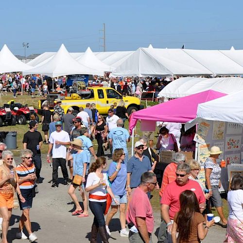 People are walking around a bustling outdoor market with many white tents and stalls displaying various items and a yellow vehicle visible.