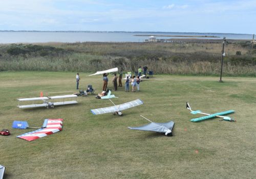 People setting up and examining model airplanes in a grassy field near water with reeds and a dock in the background. The scene is under a blue sky.