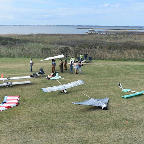 People setting up and examining model airplanes in a grassy field near water with reeds and a dock in the background. The scene is under a blue sky.