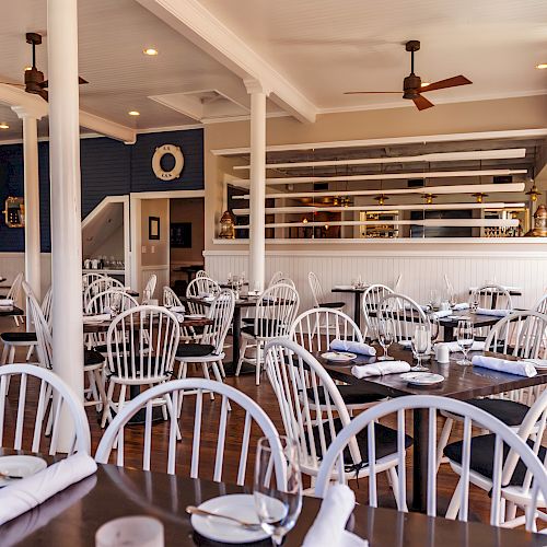 The image shows an empty restaurant with white wooden chairs and tables set for dining, adorned with napkins and plates, in a cozy, well-lit space.