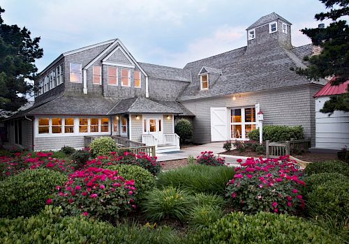 A charming house with a gray exterior, white trim, and an inviting porch is surrounded by lush greenery and blooming red flowers.