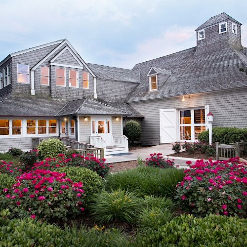 A charming house with a gray exterior, white trim, and an inviting porch is surrounded by lush greenery and blooming red flowers.