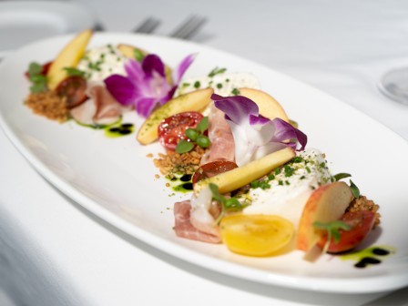 A beautifully plated dish featuring a variety of colorful ingredients, including flowers, fruits, greens, and sliced meats on a white rectangular plate.