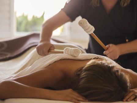 A person is receiving a back massage with wooden massage tools, while lying on a table covered with a white towel.