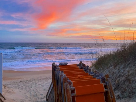 A row of orange beach chairs leads to the ocean at sunset, with a beautiful sky painted in hues of pink, blue, and purple at the Sanderling Resort.