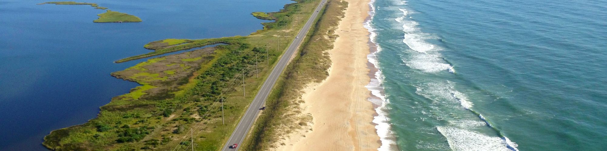 An aerial view of a coastal road running parallel to a long sandy beach and ocean waves on one side and green marshlands on the other side.