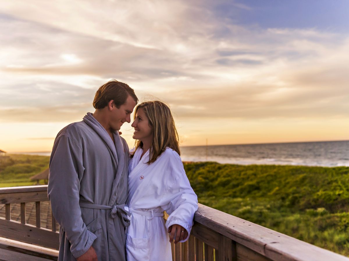 A couple in bathrobes shares a romantic moment on a wooden deck overlooking the ocean at sunset, surrounded by lush greenery and a serene sky.