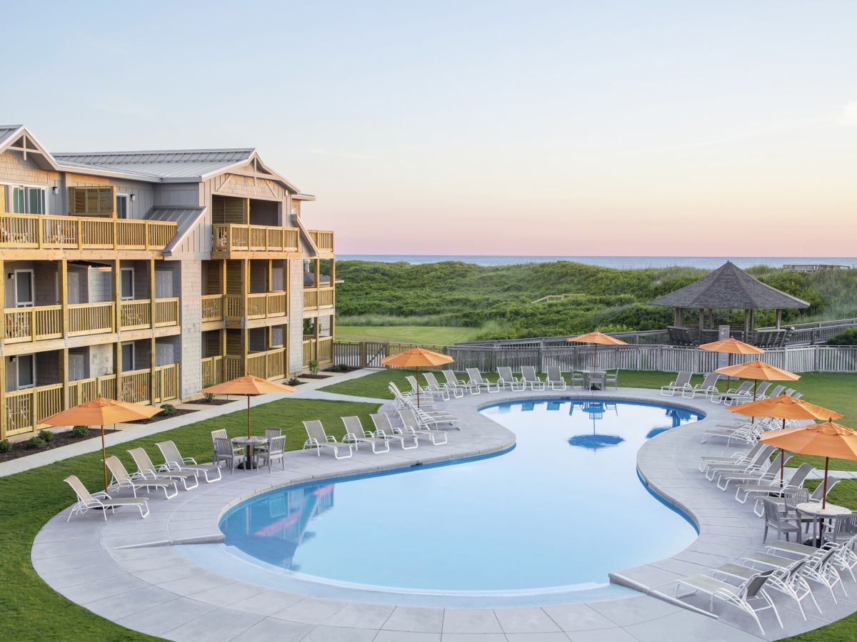 A serene beachside resort features a pool with lounge chairs and orange umbrellas, surrounded by a green lawn and overlooking sand dunes and the ocean.