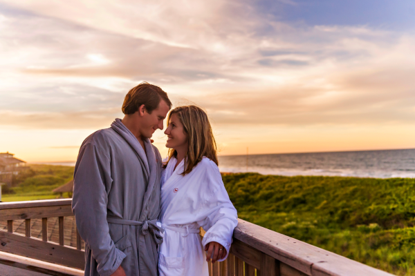 A couple in robes stands on a wooden deck overlooking the ocean, under a colorful sunset sky, sharing a close and affectionate moment together.