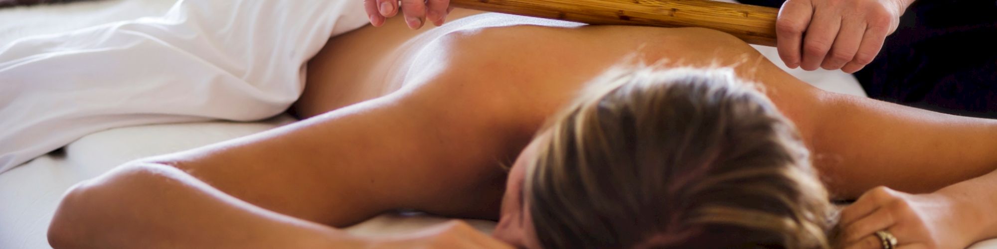 A person is lying face down on a massage table receiving a back massage with a wooden stick.