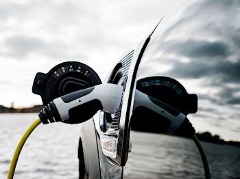 A close-up of an electric vehicle being charged with a charging plug inserted, against the backdrop of a cloudy sky and water.