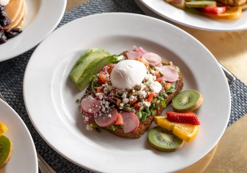 A plate of avocado toast topped with a poached egg, radishes, and various toppings, served with sliced kiwi, strawberries, and other fruits.