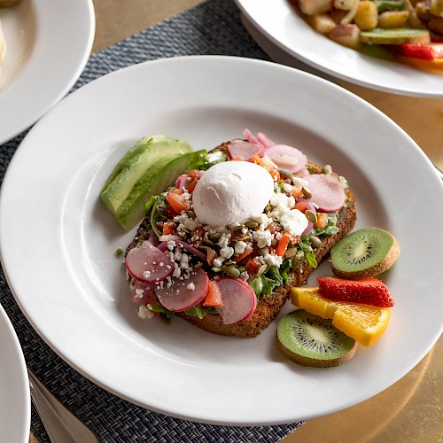 A plate of avocado toast topped with a poached egg, radishes, and various toppings, served with sliced kiwi, strawberries, and other fruits.