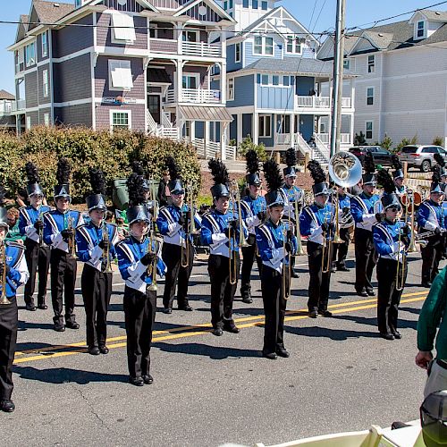 A marching band in blue and black uniforms performs in front of a row of colorful houses on a street, with people watching in the foreground.