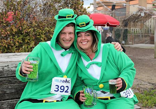 Two people dressed as leprechauns, each holding drinks, with numbers 392 and 391, sit on a bench outdoors, smiling and posing for a photo.