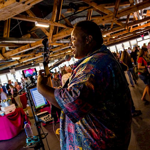 A person is playing the guitar and singing into a microphone indoors, with people socializing and mingling in the background.