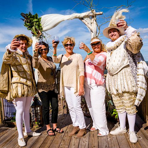 Five people stand on a wooden deck, raising glasses. Two wear historical costumes; three dress casually, with an ocean backdrop.