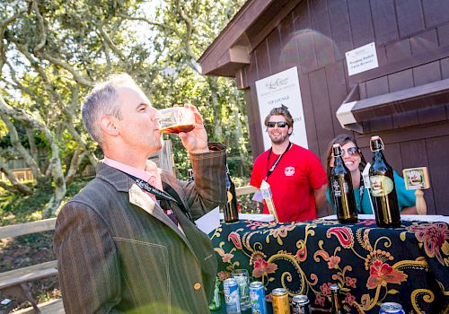 A person is drinking from a glass in front of a booth with two smiling individuals who appear to be working at a beverage stand.