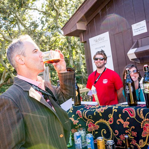 A person is drinking from a glass in front of a booth with two smiling individuals who appear to be working at a beverage stand.