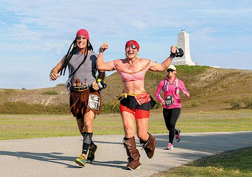 Three people are running in a race; two are dressed as pirates, while the third is in regular athletic wear with a monument in the background.