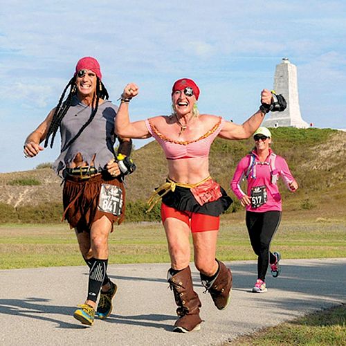 Three people are running in a race; two are dressed as pirates, while the third is in regular athletic wear with a monument in the background.