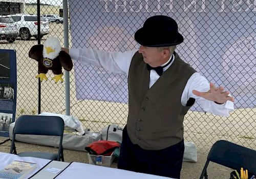 A person wearing a vest, bow tie, and bowler hat is holding a bald eagle puppet behind a table in an outdoor setting with a chain-link fence.