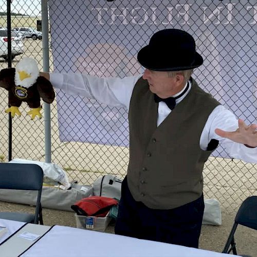 A person wearing a vest, bow tie, and bowler hat is holding a bald eagle puppet behind a table in an outdoor setting with a chain-link fence.