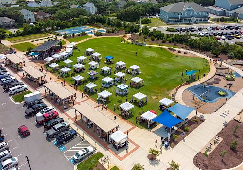 An aerial view of a community event with tents set up on a grassy area, surrounded by parked cars, walking paths, and nearby buildings.