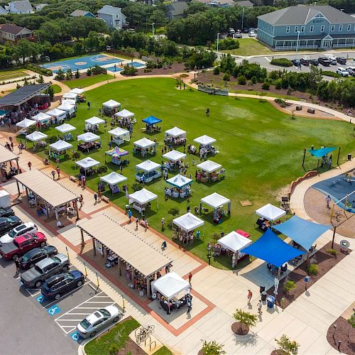 An aerial view of a community event with tents set up on a grassy area, surrounded by parked cars, walking paths, and nearby buildings.