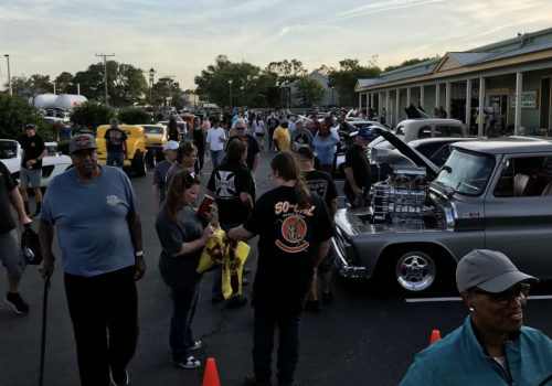 A crowd gathers at a car show in a parking lot, with various classic cars and trucks on display. People chat and stroll, enjoying the event.