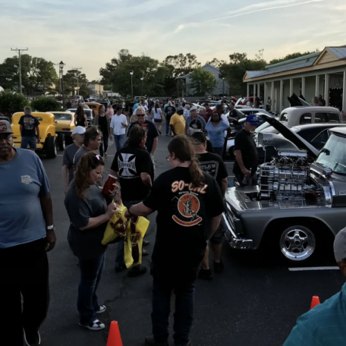 A crowd gathers at a car show in a parking lot, with various classic cars and trucks on display. People chat and stroll, enjoying the event.