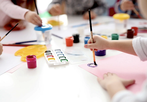Children are painting with watercolors at a table, using brushes and various colorful paints, creating artwork together in a brightly lit room.