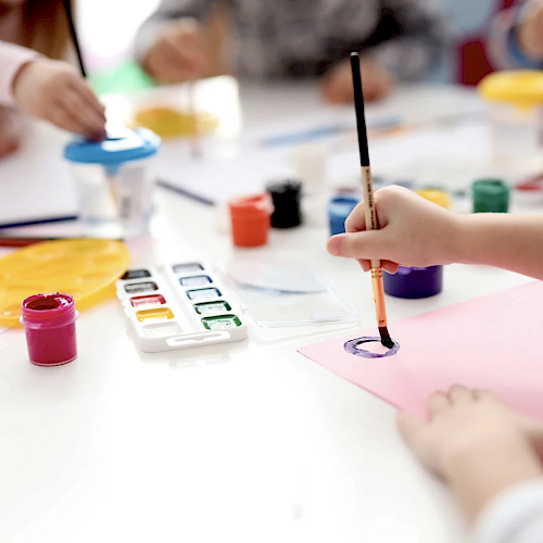 Children are painting with watercolors at a table, using brushes and various colorful paints, creating artwork together in a brightly lit room.