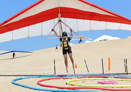 A person in a helmet and harness is hang gliding over a sandy area with colorful rings and obstacles on the ground, with a blue sky overhead.