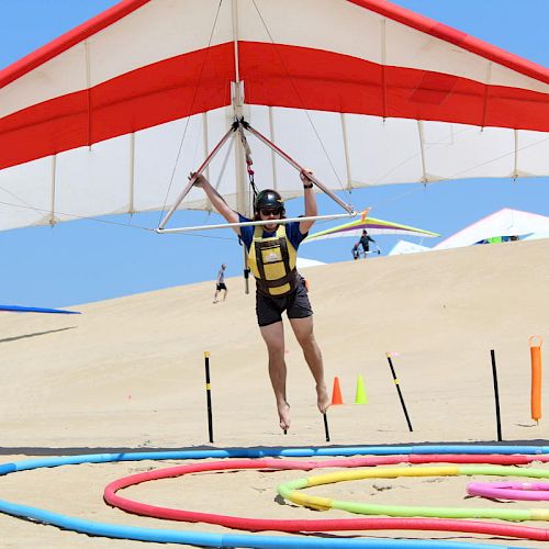 A person in a helmet and harness is hang gliding over a sandy area with colorful rings and obstacles on the ground, with a blue sky overhead.