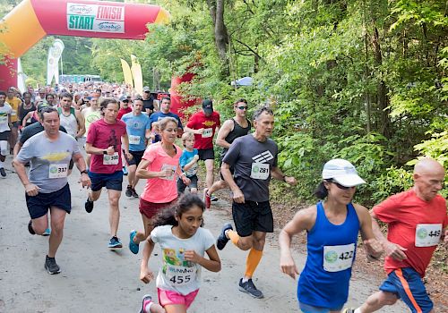 A group of people, including children and adults, are running in a race on a wooded path, passing under a large inflatable arch that says 