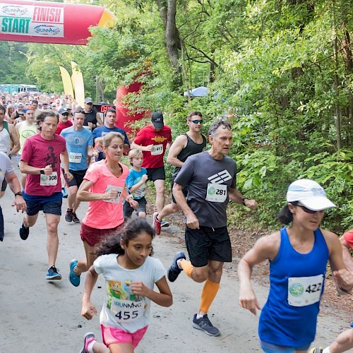 A group of people, including children and adults, are running in a race on a wooded path, passing under a large inflatable arch that says 