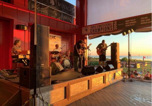 A band is performing on an outdoor stage at sunset near a coastline, under a banner reading 