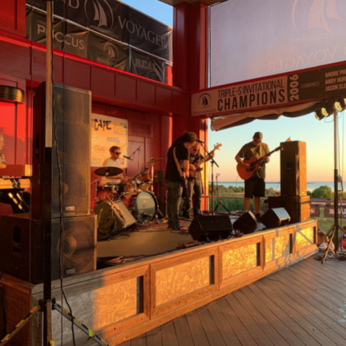 A band is performing on an outdoor stage at sunset near a coastline, under a banner reading 