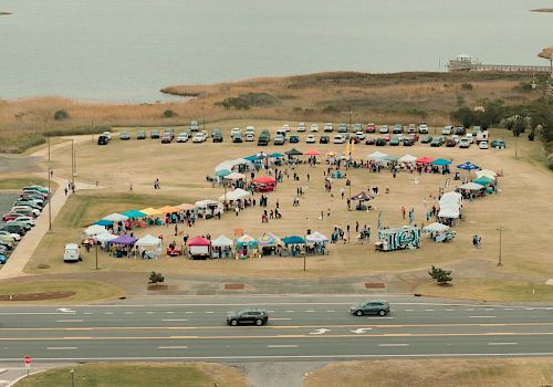 An outdoor event with numerous tents and booths arranged in a circular pattern on a grassy field near a body of water, with parked cars around it.