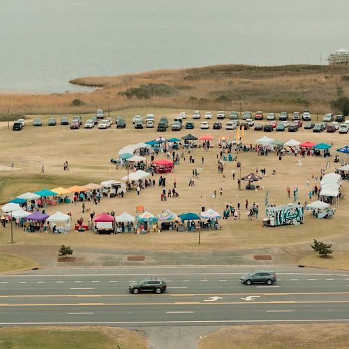 An outdoor event with numerous tents and booths arranged in a circular pattern on a grassy field near a body of water, with parked cars around it.
