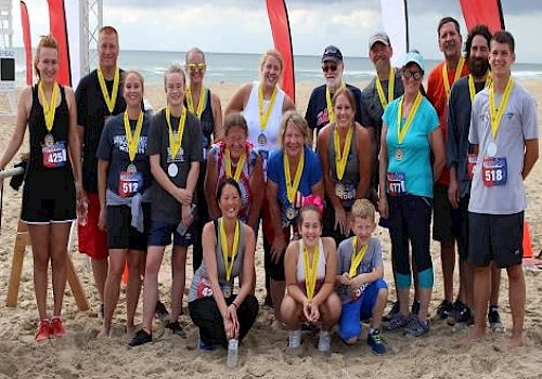A group of people with race numbers and medals posing on a beach, standing in front of a 
