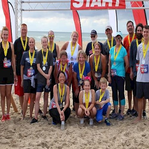 A group of people with race numbers and medals posing on a beach, standing in front of a 