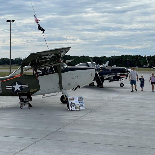 People are walking near military aircraft displayed on a tarmac, with informational signs placed in front of the planes.