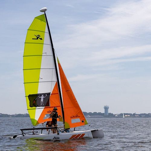 The image shows a sailboat with colorful sails on a body of water, with a couple of people onboard, and a distant shoreline with buildings in the background.