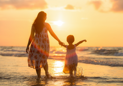 A woman and a child are walking hand-in-hand along the shore at sunset, with the ocean waves gently touching their feet.