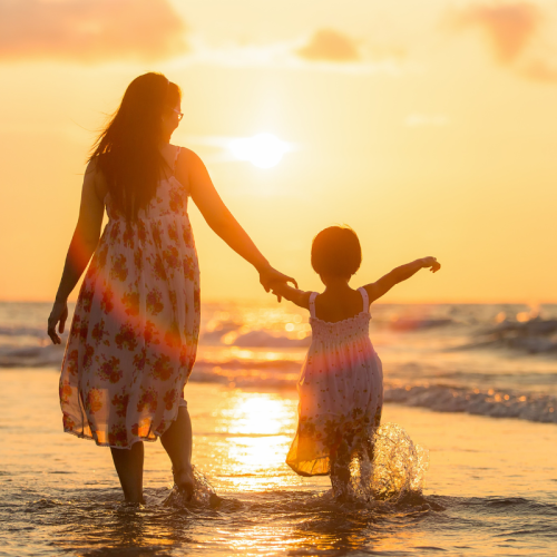 A woman and a child are walking hand-in-hand along the shore at sunset, with the ocean waves gently touching their feet.