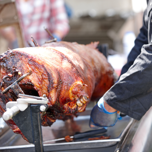 The image shows a roasted whole pig on a spit with people in the background, likely at an outdoor event or a barbecue setting.